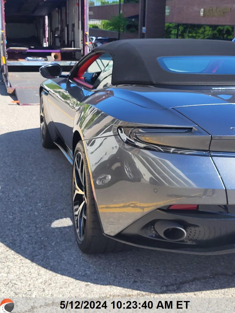 Sleek metallic gray convertible Aston Martin with red interior, ready for enclosed auto transport with a luxury car hauler in the background during vehicle shipping process