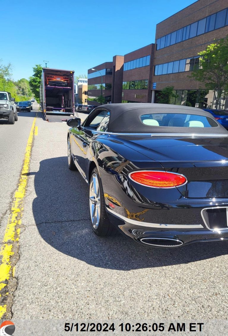 Black Bentley luxury convertible sports car parked on the street, ready for enclosed auto transport with a vehicle hauler in the background, during a car shipping process.