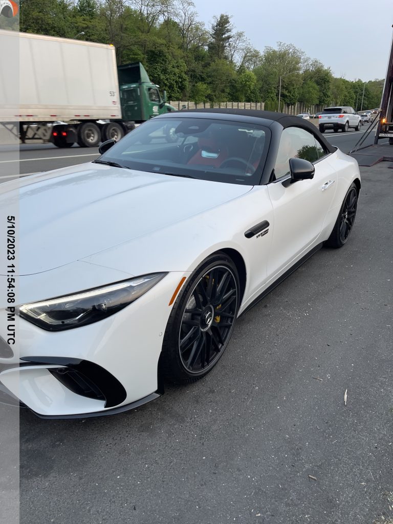 White convertible Mercedes-Benz sports car with black rims, staged for auto transport alongside a highway with trucks and cars passing by in the background, ready for luxury vehicle shipping